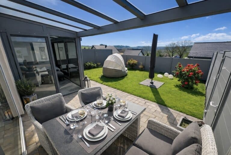 Überdachte Terrasse mit gedecktem Tisch und Sitzmöbeln, angrenzender Garten mit Liegeinsel, Sonnenschirm und blühenden Pflanzen